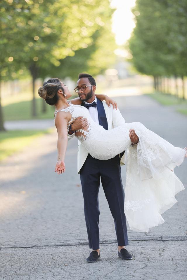Groom is carrying bride in white wedding dress at park alley